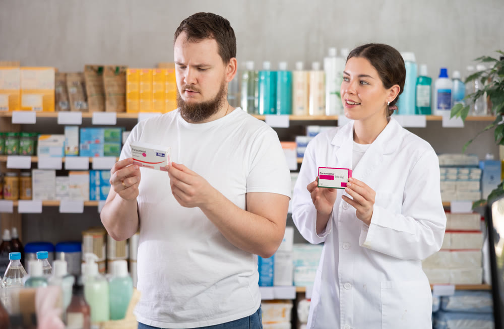 Man chooses paracetamol pills in modern pharmacy. Female pharmacist helps with choosing medicine
