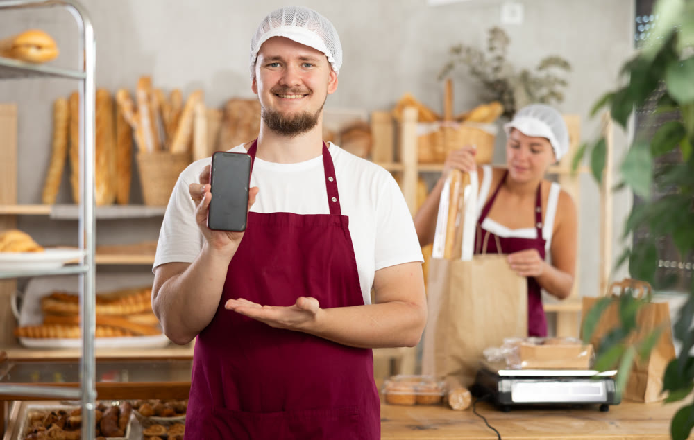Guy seller in bakery hold out hand with phone, points at screen with finger