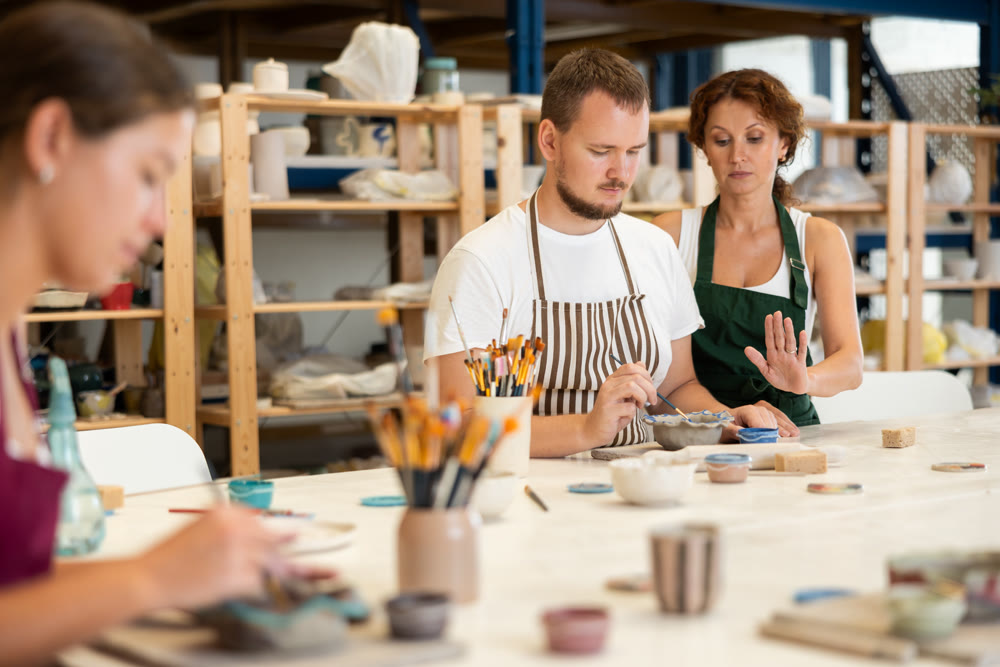 During master class, student paints clay vessel under guidance of female teacher.