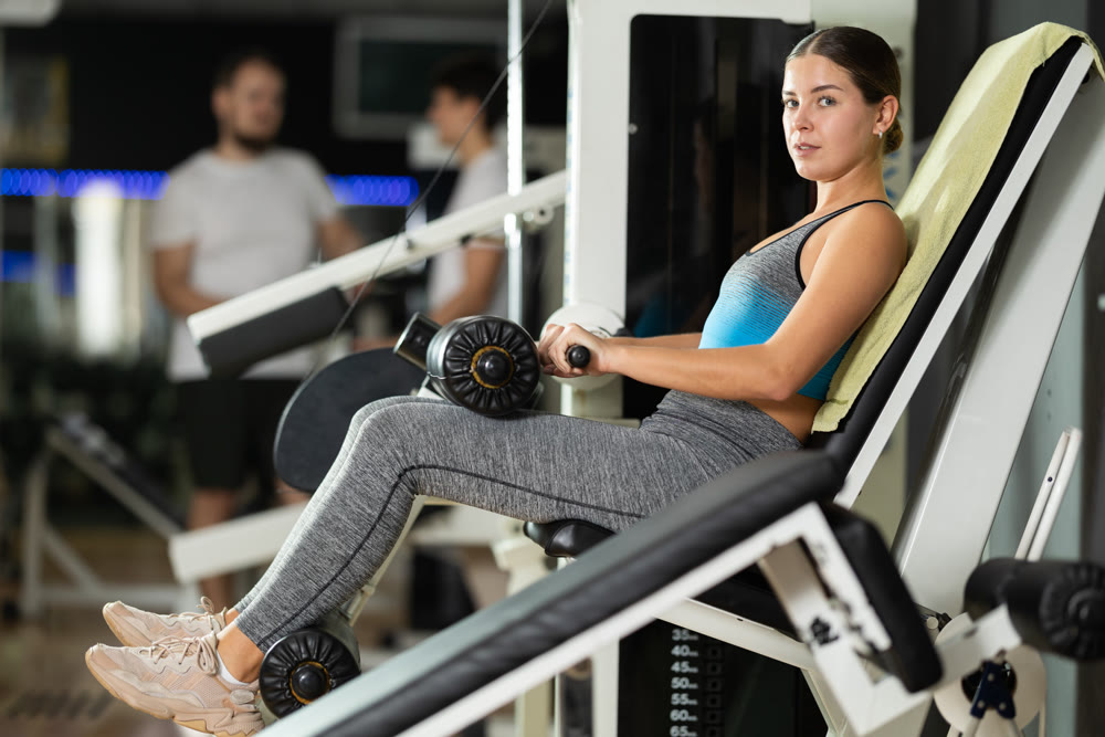 Young woman in gym performs exercises for leg muscles using simulators and fitness center equipment.
