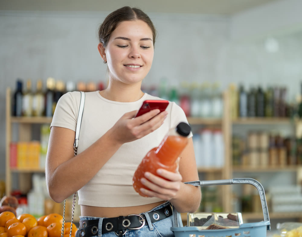 European young woman customer at shop scans QR code on grapefruit juice bottle using phone camera