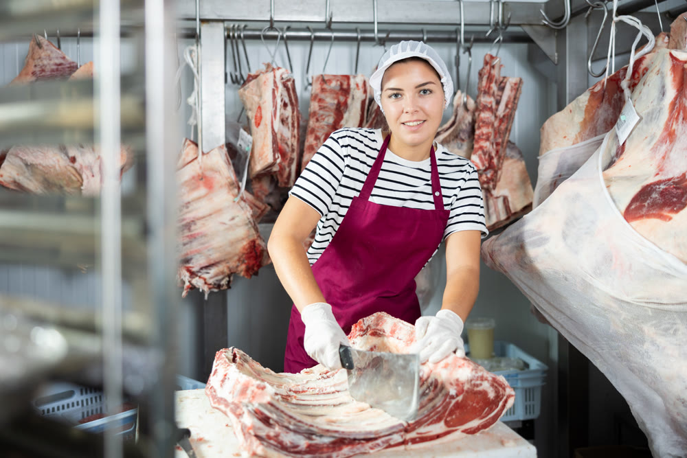 Female employee of local butcher shop divides large huge piece of meat with knife