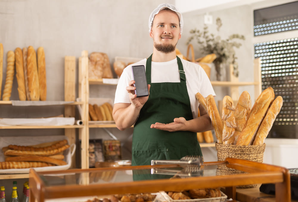 Guy seller in bakery hold out hand with phone, points at screen with finger
