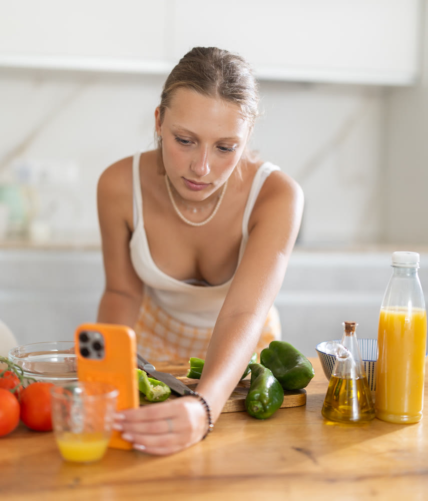 Young girl looking at smartphone while preparing fresh salad in kitchen