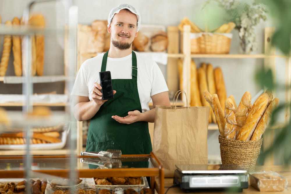 Young male bakery seller showing information on phone