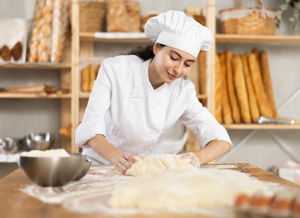 Young female baker kneading dough on table