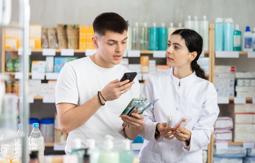 Positive young guy buyer at pharmacy choosing best toothbrush, scan QR code, young girl pharmacist consulting and offering variety of goods to customer