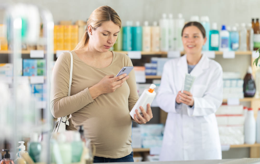 Pregnant woman in pharmacy scans QR code on antifungal cream, pharmacist offer similar product in background.