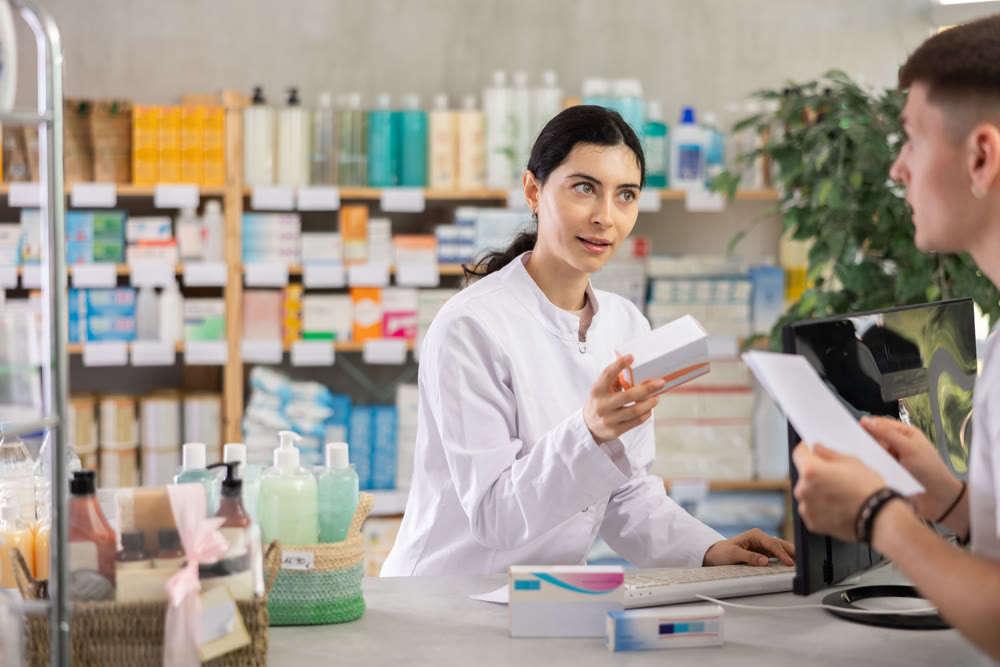 Young professional female parmacist working computer and helping young guy with prescription in hand look for medicine in drugstore