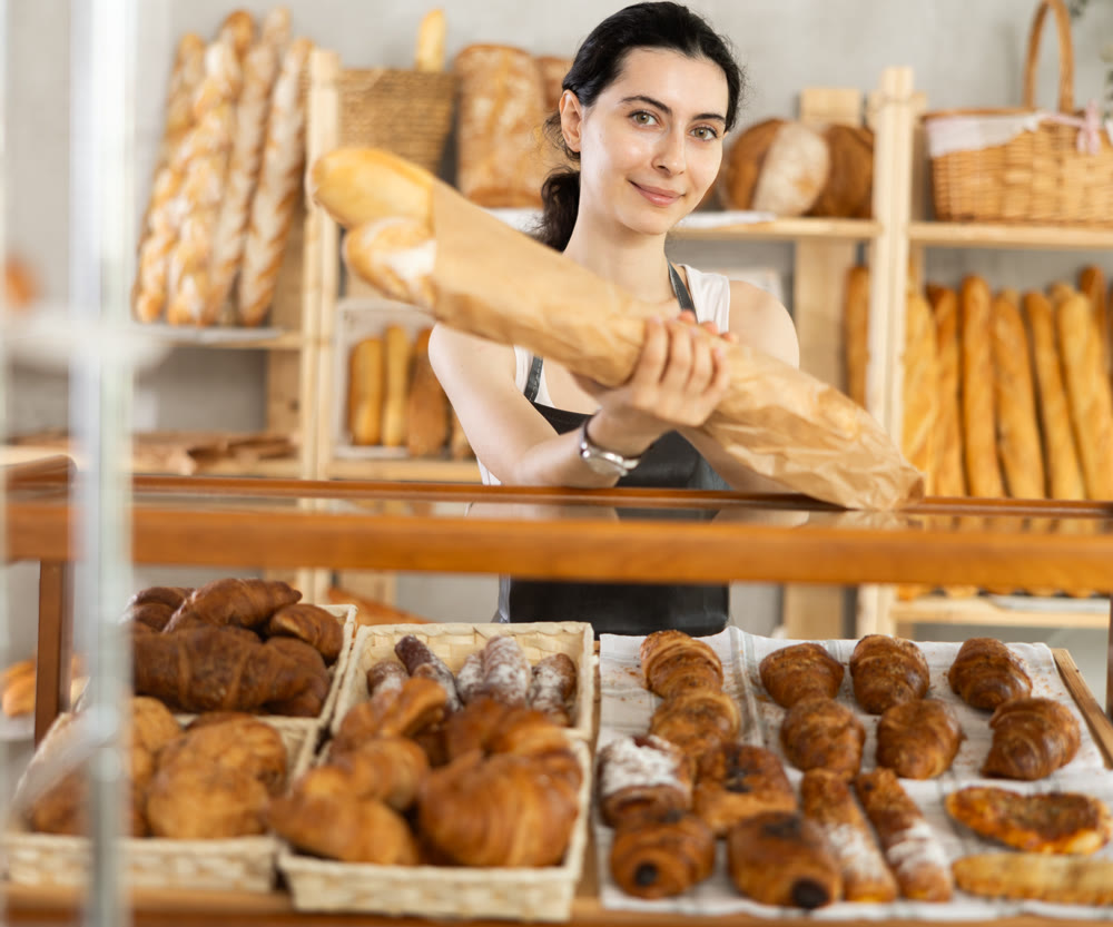 Young woman seller took fresh bagguetes of white flour out of window in bakery