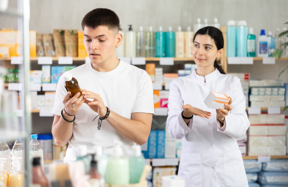 Young man buyer choosing medicinal liquid syrup standing between shelves, professional pharmacist woman helping and offering supplements at pharmacy