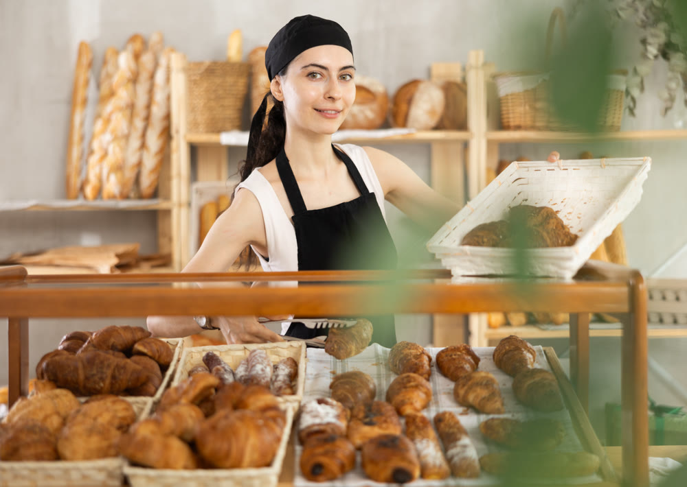 Young woman employee puts croissants in window, arranges display of goods at bakery.