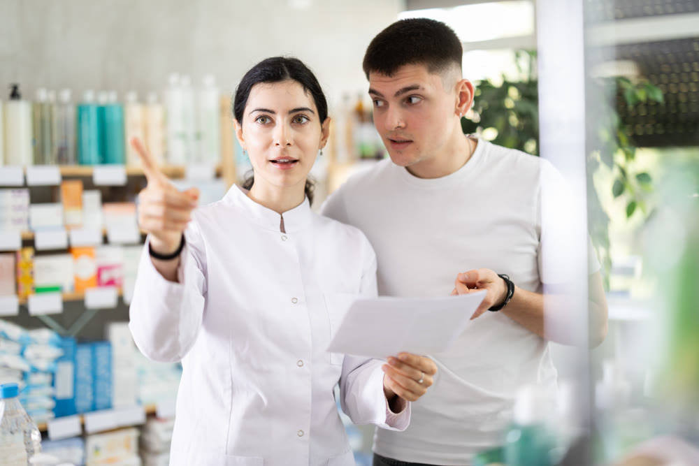 Young guy with medical prescription in hand looking for something in drugstore, young girl pharmacist consulting and helping client