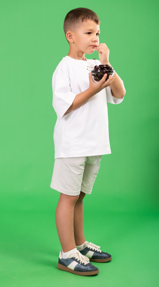 Boy in white t-shirt and shorts happily eats ripe cherries on green background