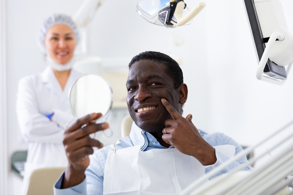 Satisfied patient examines cured teeth using mirror while sitting in dentist chair