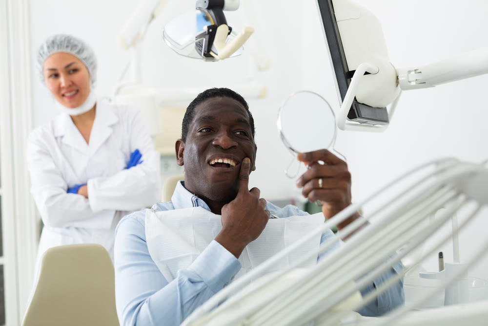 Satisfied patient examines cured teeth using mirror while sitting in dentist chair