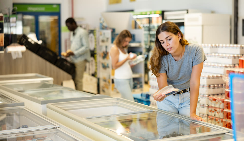 Female shopper carefully chooses frozen food in grocery supermarket