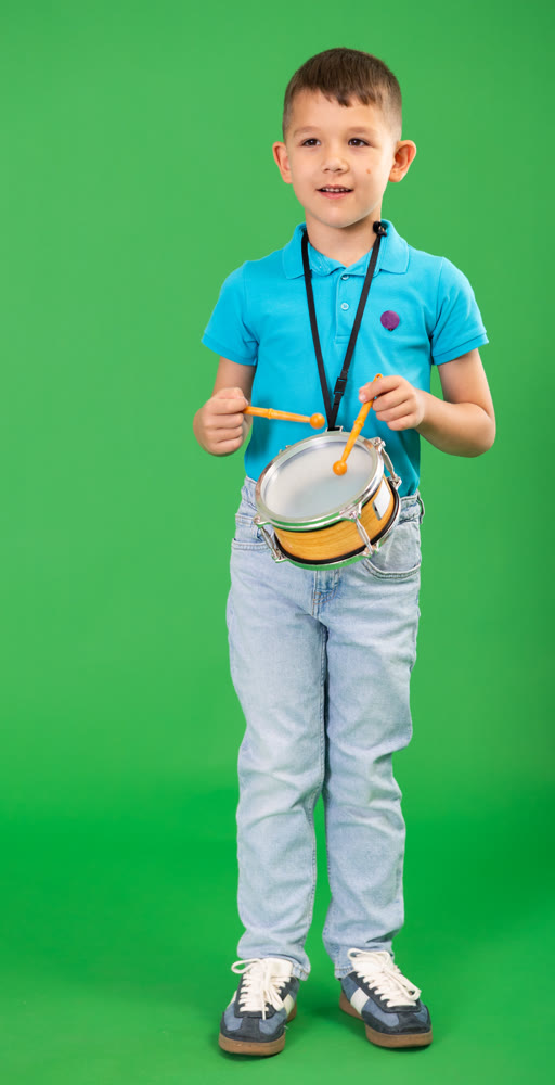 Boy in blue jeans drums on children drum that hangs from his neck against green background