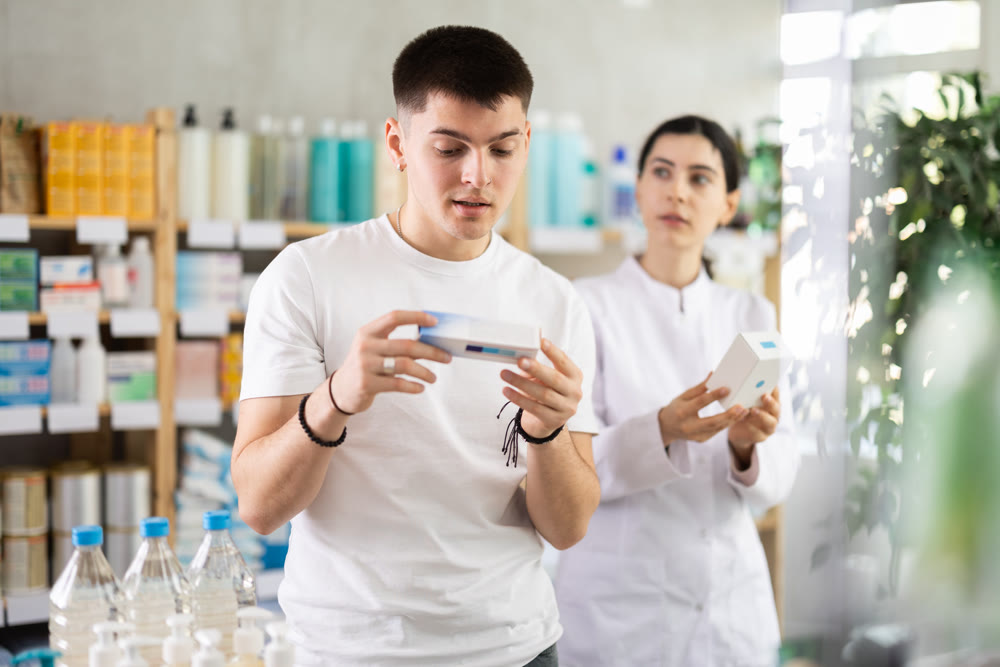 Young female pharmacist helping young man choosing skin care lotion or cream in drugstore