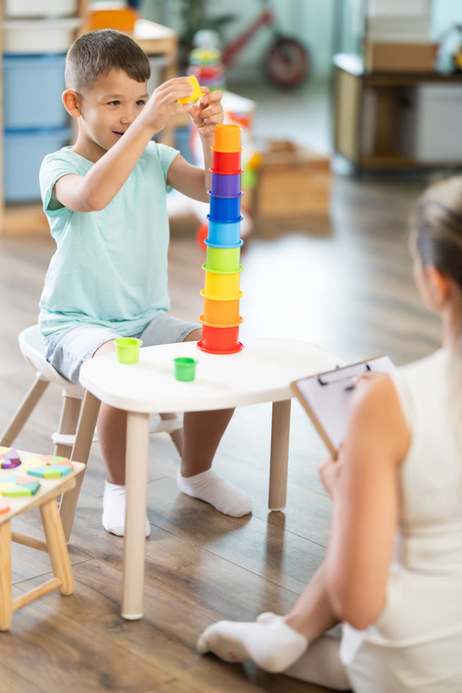 Boy plays with multi colored plastic cups, placing them inside each other. Psychologist helps and observes.
