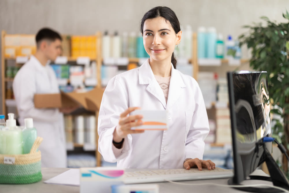 Portrait of young woman pharmacist standing at prescription counter and working on computer, stocktaking medicines at pharmacy shop and male assistant in background