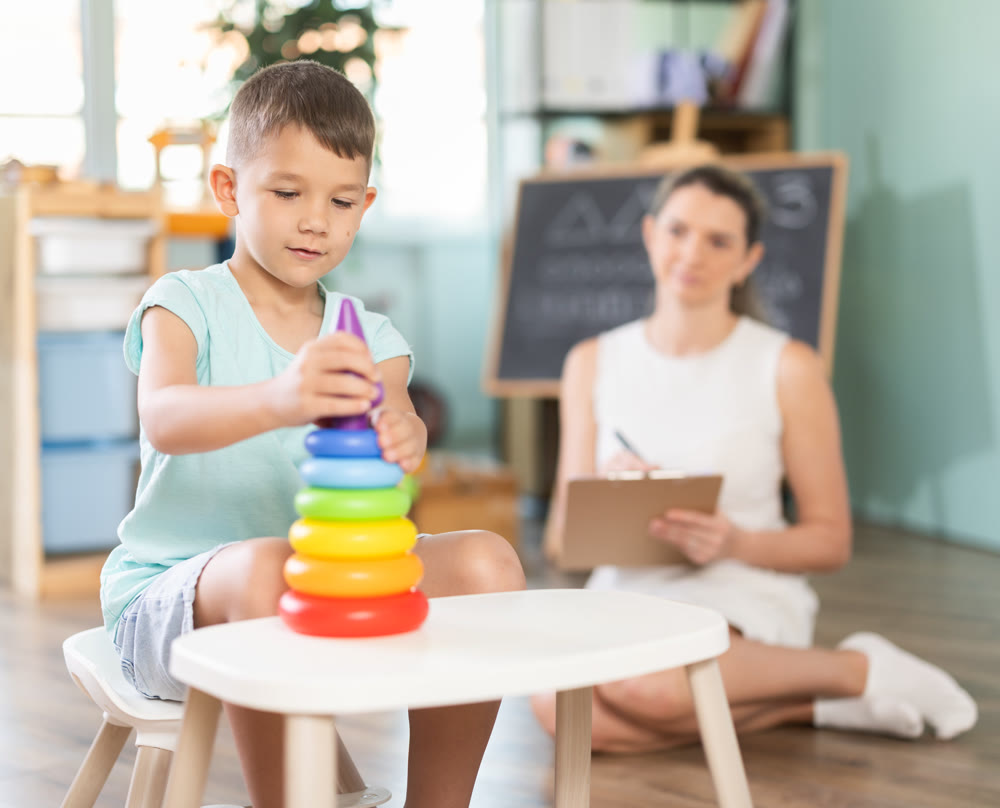 Little boy assembling toy pyramid with psychologist observing in background