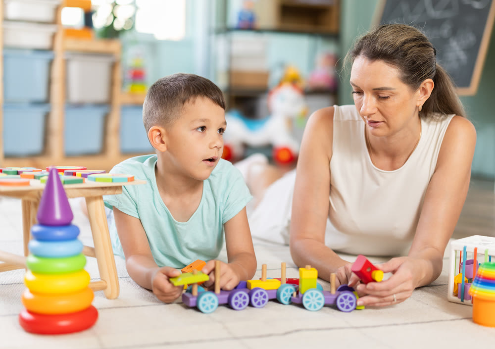 Little boy assembling toy pyramid on the floor with psychologist in background