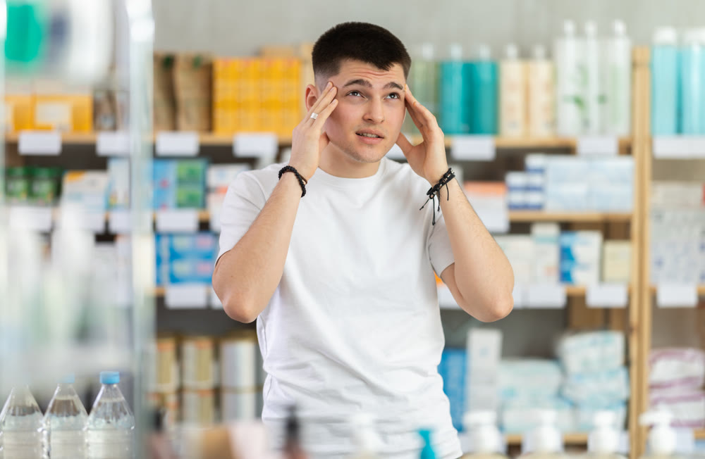 Sick man standing between shelves in drugstore suffering from headache or having migraine, choosing medicine. Health care and medicine concept