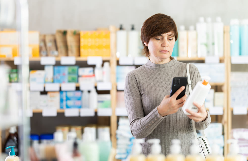 Female customer using smartphone while choosing skincare product in pharmacy