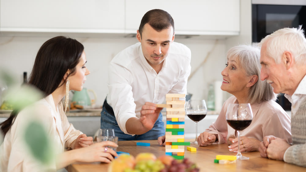 Joyful senior parents and young couple sitting around table playing brick game