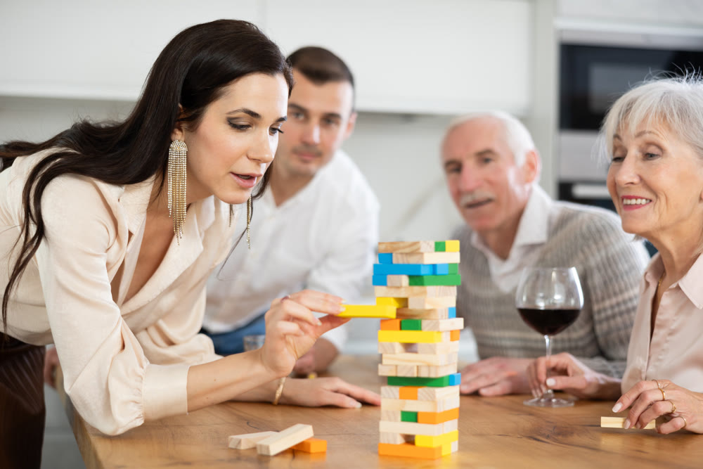 Group of mature spouses and son with girlfriend is playing board game with wooden blocks in kitchen