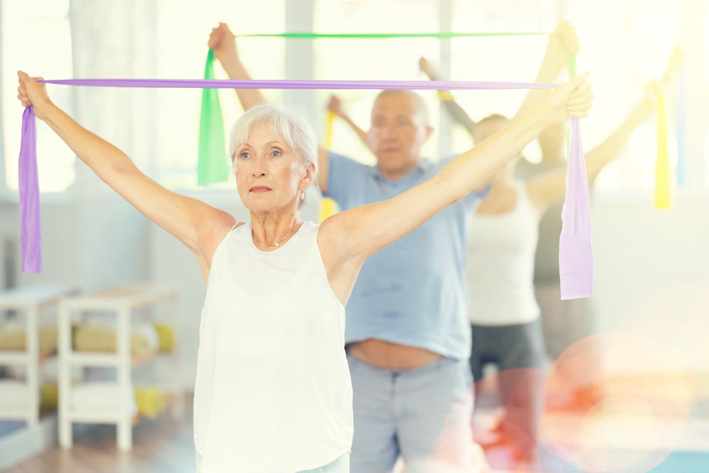 Elderly woman kneeling performs exercises with wide elastic band during Pilates workout.
