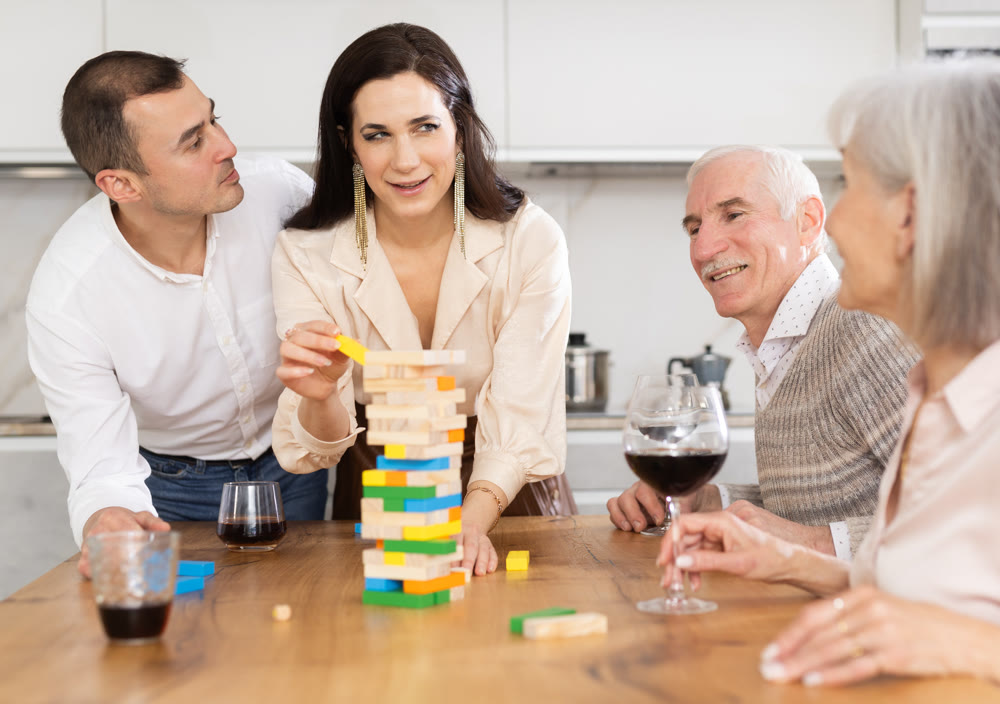 Joyful senior parents and young couple sitting around table playing brick game