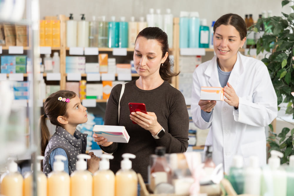 Woman with daughter in pharmacy scans QR code on tablet box, pharmacist in background