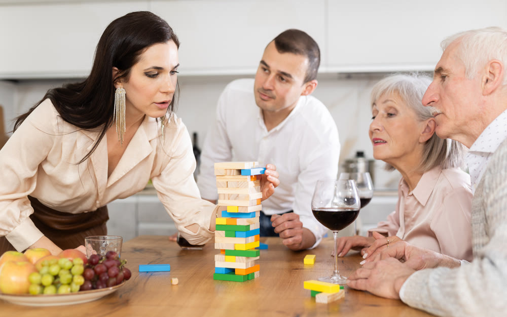 Group of mature spouses and son with girlfriend is playing board game with wooden blocks in kitchen