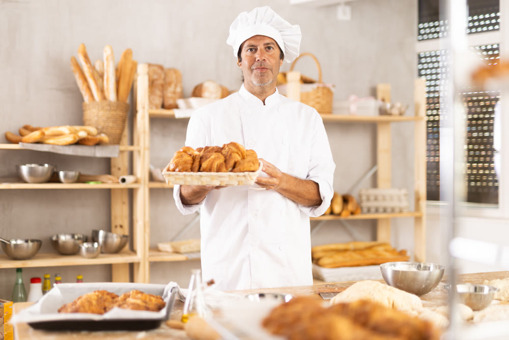 Caucasian male cook-baker worker with basket of croissant, shows many products