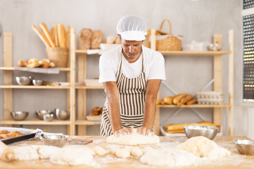 Caucasian guy works in bakery as baker, kneads dough, works with flour.