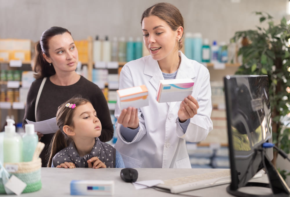 Young female pharmacist serving mother with daughter standing by computer