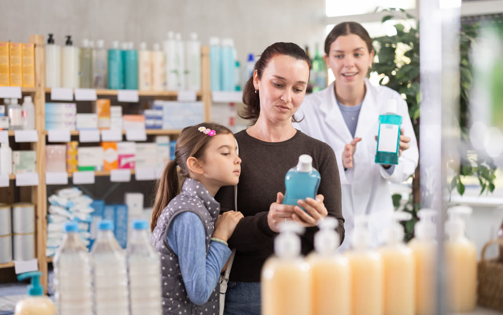 Woman with schoolgirl daughter are busy choosing mouthwash, pharmacist offer help in background