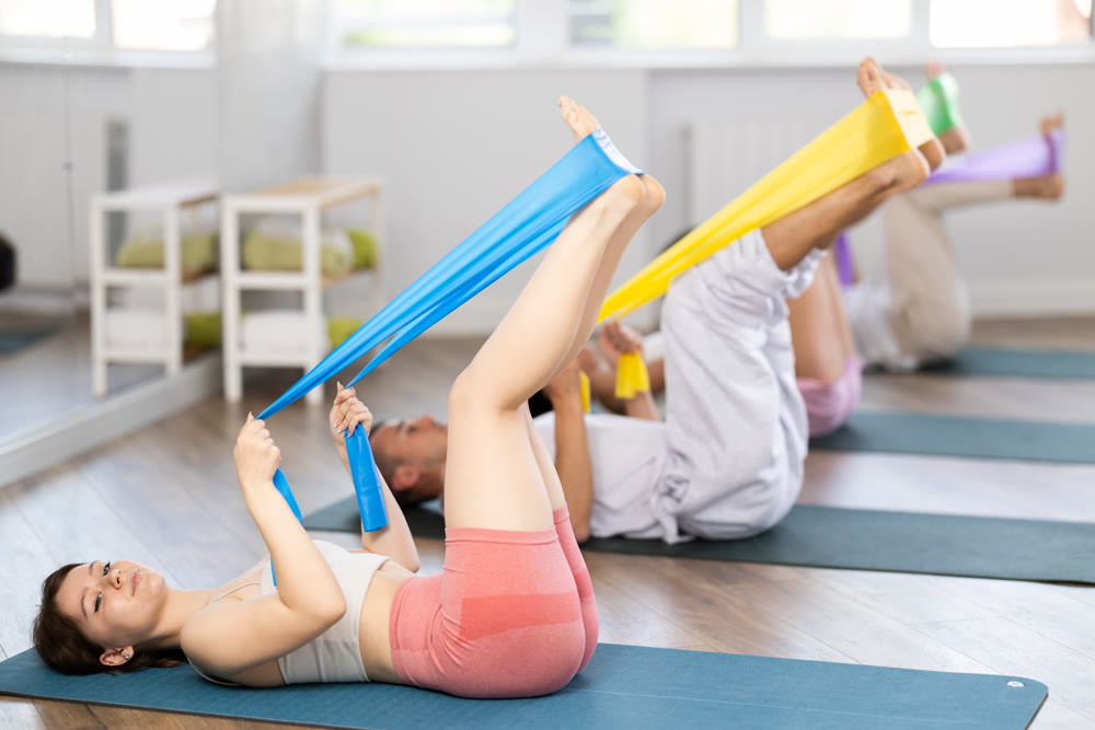 Sports girl with women and men of different ages doing stretch exercises for legs with resistance bands while lying on mats in gym