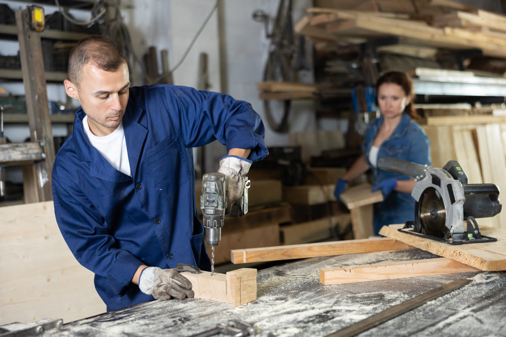 Male employee of carpentry workshop uses drill to make holes in wooden billet