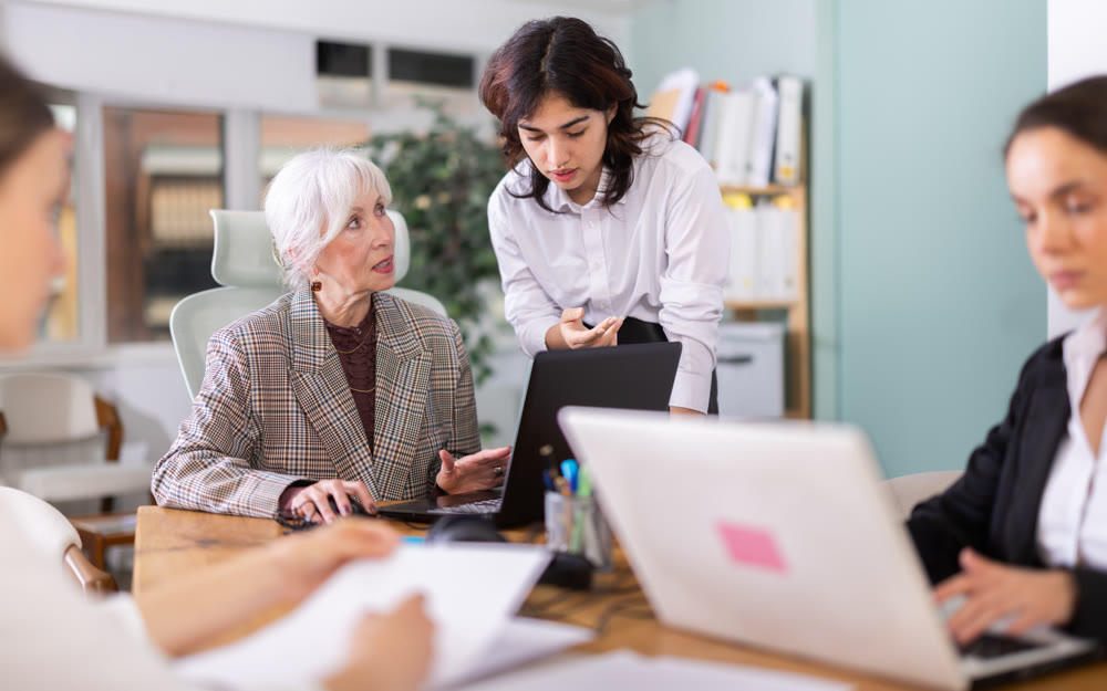 Asian girl leaned over to seated senior woman employee, helps with work and looks at laptop screen