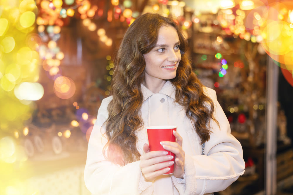 Smiling woman walking through Christmas street market with warm beverage in paper cup