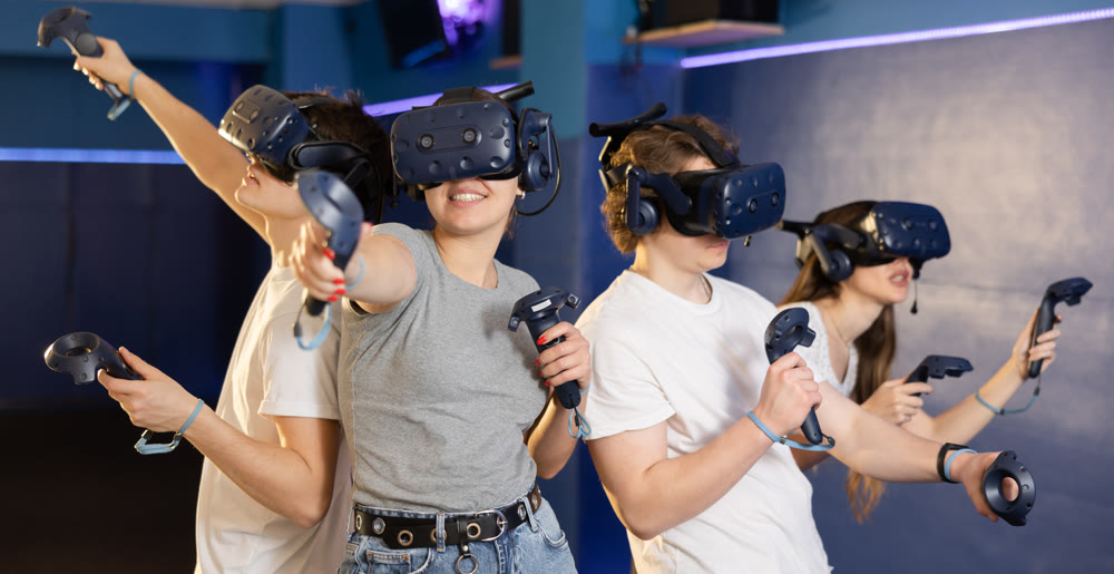 Young people in virtual reality glasses stand back to back and play game by controlling joysticks