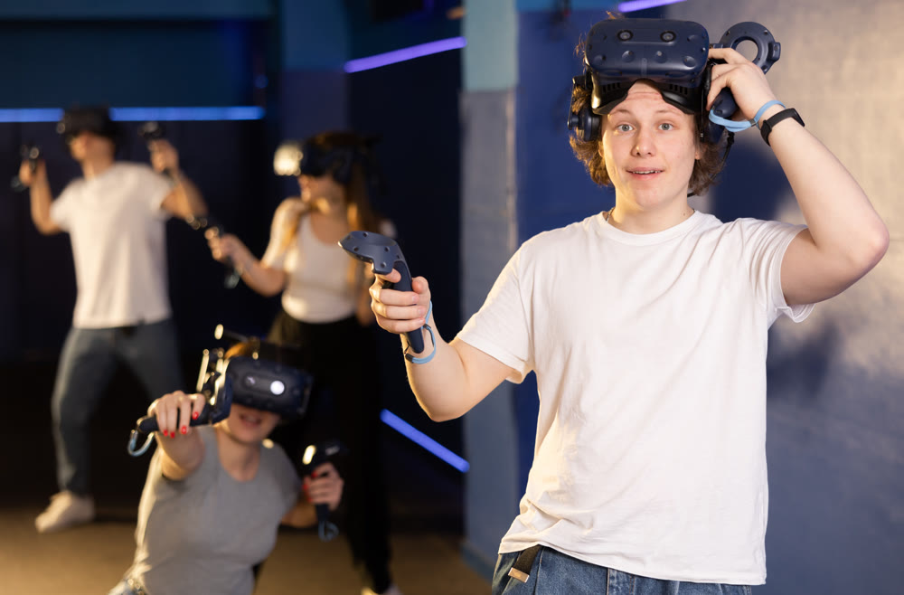 Emotional young guy in VR headset with joysticks playing with friends