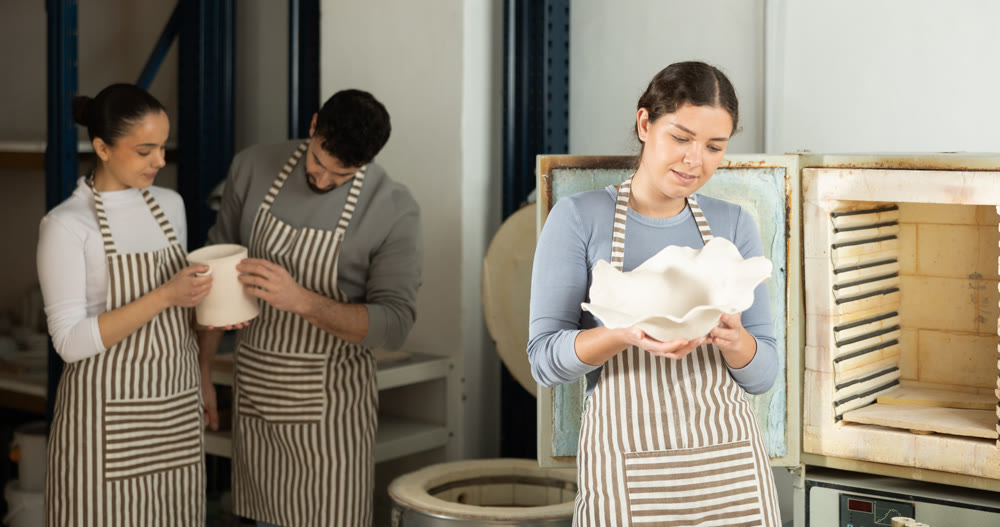 Two female potters take finished ceramic dishes, various mugs and a vase out of oven