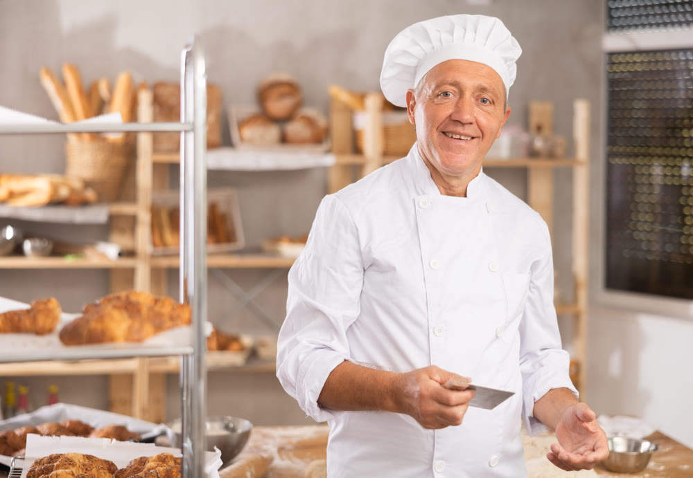 Mature man baker in white coat overall and cap stands in kitchen of bakery
