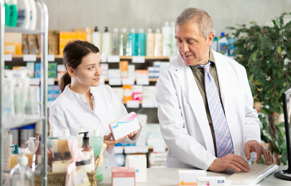 Elderly experienced pharmacist works at computer in drugstore. Young assistant helps to find pills