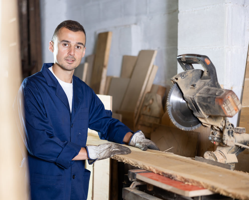 Male employee of workshop cuts wooden board into narrow parts using circular saw