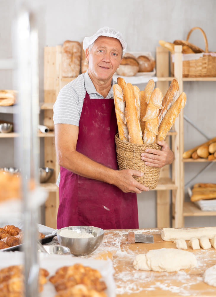 Smiling elderly baker presenting wicker basket of fresh baguettes